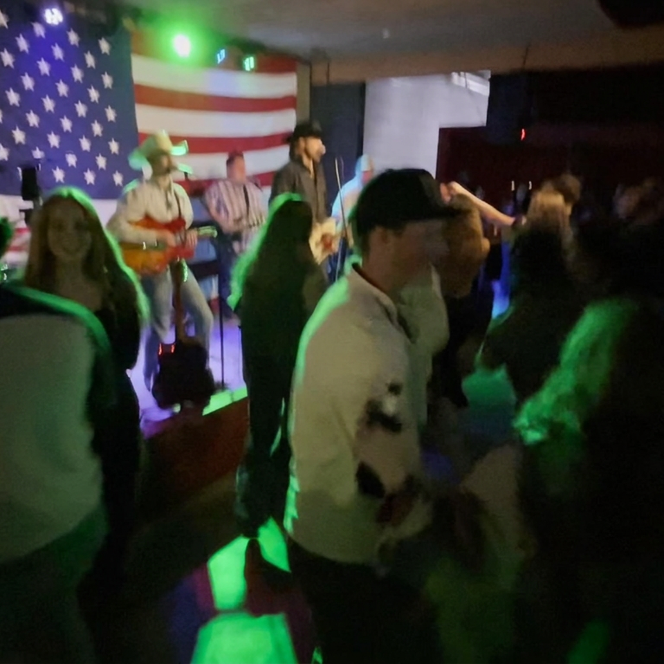 Crowd dancing before a live band performing under colorful lights with an American flag backdrop.