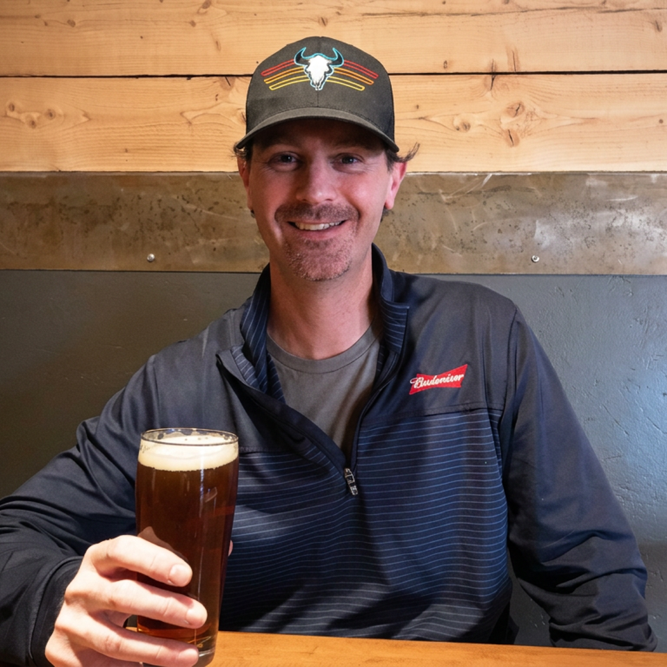Smiling restaurant staff member in a branded cap and jacket holds a pint of dark beer against a rustic wood and metal wall.